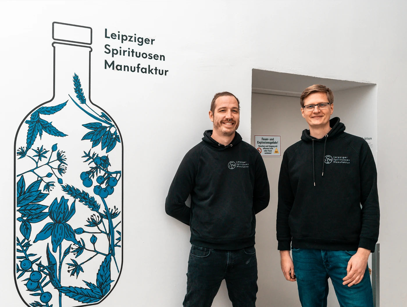 Jan Enno Cöster und Peter Morczinek vor dem Eingang der Produktion der Leipziger Spirituosen Manufaktur neben dem Keyvisual